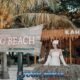 girl in white dress standing on beach next to large wooden sign for kahung beach in front of bar